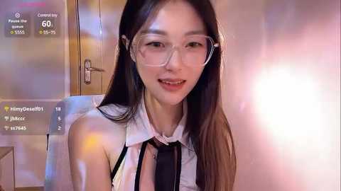 Media: Video of an Asian woman with long black hair, wearing glasses, a white sleeveless top, and a black tie, smiling. Background shows a blurred, warm-toned room with a wooden door and a bathroom light.