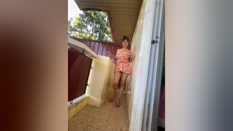 Media: Video of a young Asian woman with light skin and medium-length dark hair, wearing a pink floral dress, standing on a balcony with beige floor tiles, maroon wall, and a green tree background.