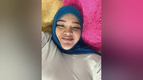 Media: Video of a smiling, light-skinned woman with brown eyes, wearing a turquoise hijab and a beige shirt, lying on a pink and yellow fuzzy blanket.