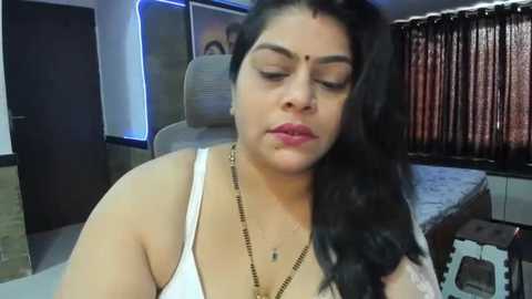 Media: A video of a South Asian woman with long black hair and fair skin, wearing a white tank top and gold necklace, sitting on a chair in a dimly lit bedroom with brown curtains.