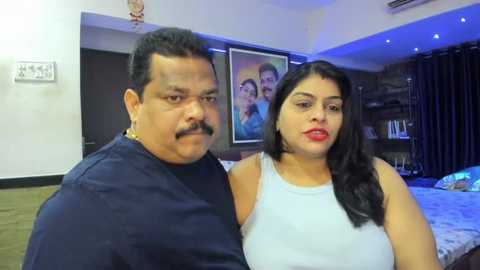 Media: Video of an overweight Indian man with a mustache and a woman with long black hair in a white sleeveless top, standing in a modern, dimly lit living room with a framed picture of a family behind them.