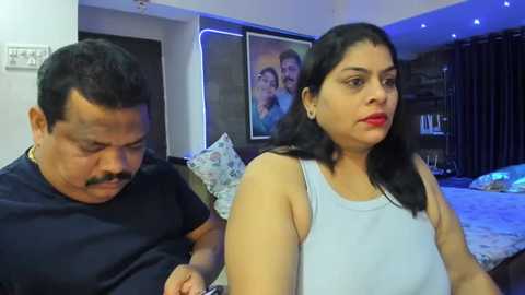 Media: Video of an Indian couple in a modern living room; man with a mustache, woman with long black hair and red lipstick, both in casual attire.