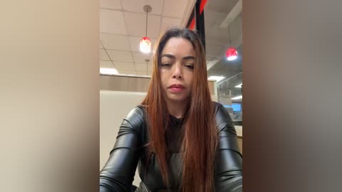 Media: Video of a Latina woman with long, straight, auburn hair, wearing a black leather jacket, sitting indoors with fluorescent lights and red pendant lights.