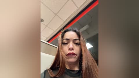 Media: Video of a Latina woman with long, straight brown hair, wearing a black shirt, in a bathroom with beige walls and a red-framed mirror. She has a neutral expression, looking downwards.