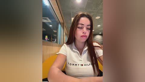 Media: Video of a young woman with long brown hair, fair skin, wearing a white Tommy Hilfiger polo shirt, sitting in a modern restaurant with wooden walls and large windows.