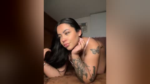 Media: Video of a young woman with long black hair, medium skin tone, and tattoos on her arms, lying on her stomach in a bedroom, wearing pink lingerie.