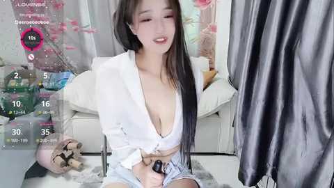 Media: A young Asian woman with long black hair, wearing a low-cut white blouse, sits on a white sofa, holding a black vibrator, in a modern, light-colored room.