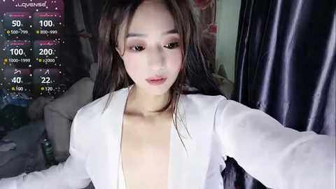 Media: Video of an Asian woman with long black hair, wearing a white blazer, in a dimly lit room with a bed, a TV, and a dark curtain in the background.