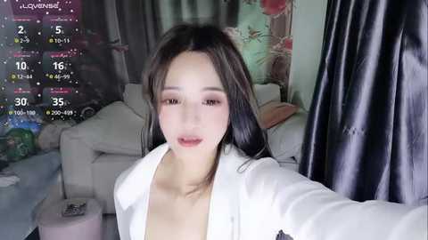 Media: Video of a young Asian woman with long black hair, fair skin, and small breasts, wearing a white jacket, in a messy living room with a calendar display.