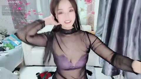 Media: Video of an Asian woman with long black hair, wearing a sheer black top, posing in a living room with white furniture and pink flowers.