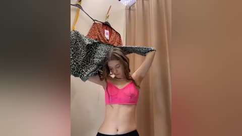 Media: A video of a young woman with fair skin, brown hair, and slender physique, wearing a pink lace bra and black pants, adjusting a leopard-print dress in a beige-walled changing room.