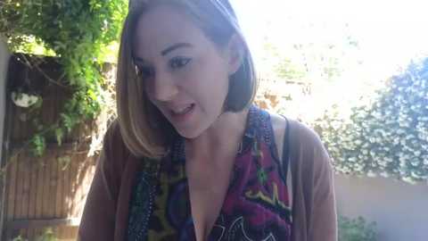 Media: A video of a smiling young woman with shoulder-length blonde hair, wearing a colorful, patterned vest over a brown top, standing outdoors in a sunny garden with greenery and a wooden fence in the background.