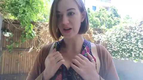 Media: Video of a young woman with light skin, brown hair, and a floral top, pulling up her shirt in a sunlit backyard.