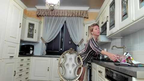 Media: A video of a blonde woman in a striped dress washing dishes in a quaint, vintage kitchen with cream-colored cabinets, tiled backsplash, and a window with lace curtains.