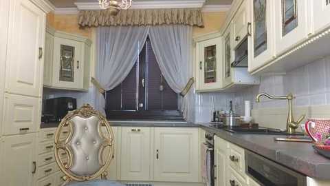 Media: A video of a vintage-style kitchen with cream cabinets, a tufted chair, and a window with sheer curtains. The countertops are black, and the backsplash is tiled in white.