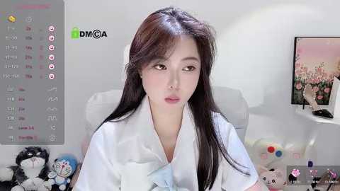 Media: Video of an East Asian woman with long black hair, wearing a white shirt, sitting in a white chair, surrounded by plush toys and a TV in a minimalist room.