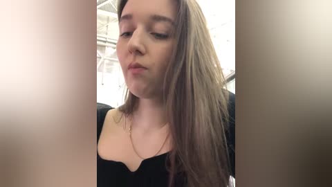 Media: Video of a young Caucasian woman with long, straight brown hair, wearing a black top, looking down introspectively. The background shows an industrial setting with metal structures and white walls.