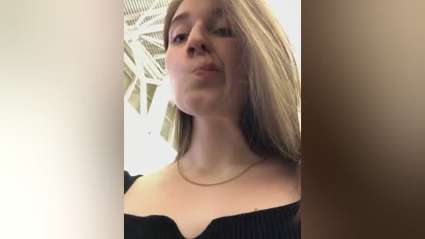 Media: Video of a young Caucasian woman with fair skin, long blonde hair, wearing a black off-shoulder top. She has a neutral expression. Background shows a modern, geometric ceiling pattern.