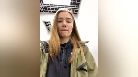 Media: Video of a young woman with long, straight blonde hair, wearing a green jacket over a dark shirt, standing in a modern, white-walled room with grid-patterned ceiling tiles.