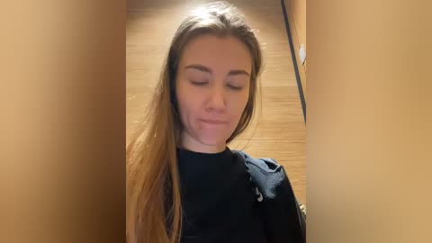 Media: Video of a young Caucasian woman with long, straight, light brown hair, wearing a black shirt, looking downward with a slight smile. Background shows wooden floor and beige walls.