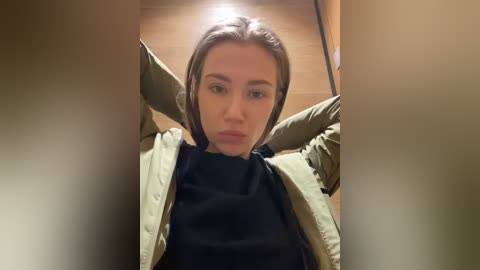 Media: A video of a young woman with straight, brown hair, wearing a black turtleneck under a green jacket, arms raised, in a dimly lit room with wooden flooring.