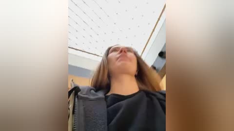 Media: A video of a young woman with long brown hair, wearing a black shirt, standing in a modern, well-lit room with a grid-patterned ceiling.