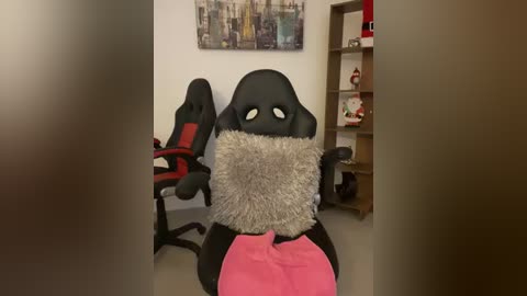 Media: Video of a plush, black, pillow-faced chair with a gray shaggy cushion and pink blanket. Background features a modern cityscape painting, red gaming chair, bookshelf, and toys.