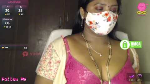 Media: A video of a South Asian woman in a floral pink bra, floral face mask, and a floral dupatta, sitting in a dimly lit room with a TV displaying a live stream.