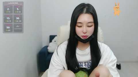 Media: Video of an Asian woman with long black hair, wearing a white T-shirt, black mask, and green jacket, seated in a simple room with a digital clock and a stuffed toy.