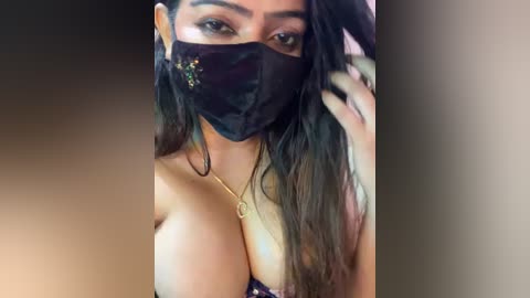 Media: Video of a woman with long dark hair, wearing a black face mask and a necklace, her large breasts partially visible.