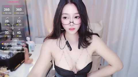 Media: A video of an East Asian woman with long black hair, wearing glasses, a black strapless top, and a necklace, seated in a dimly lit room with a computer monitor displaying social media statistics.