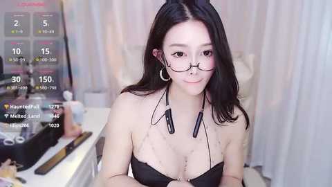 Media: Video of an East Asian woman with long black hair, wearing glasses, a black strapless top, and headphones. She sits at a desk with a gaming setup, including a monitor displaying game stats.