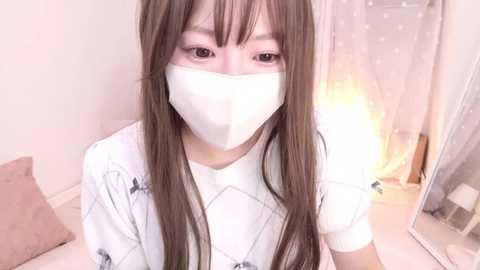 Media: Video of an Asian woman with long brown hair and pale skin, wearing a white mask, white shirt, and white patterned vest, sitting in a softly lit room with a mirror and white curtains.