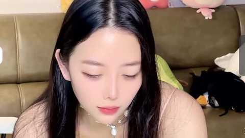 Media: Video of a young East Asian woman with long, straight black hair, light skin, and closed eyes, wearing a beige top, sitting on a brown couch, with a baby on the backrest.