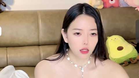 Media: Video of an Asian woman with long black hair, light skin, and full lips, wearing a pearl necklace, sitting on a beige couch with stuffed animals.