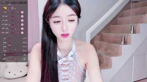 Media: Video of a young East Asian woman with long black hair, wearing a revealing, white lace top, sitting on a beige couch in a modern, minimalist living room with a staircase in the background.