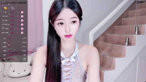 Video of a young Asian woman with long black hair, wearing a white lace-up top, sitting on a staircase. The background features a beige teddy bear and a pink wall.