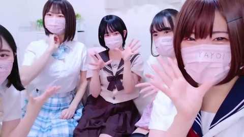 Media: Video of five Japanese schoolgirls in traditional sailor uniforms, wearing face masks, waving, smiling, and seated on a bed.