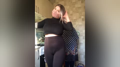 Media: A video of a plus-size woman with fair skin, wearing a black crop top and high-waisted leggings, smiling with a pink mask on, in a kitchen with beige tiles and stainless steel appliances.