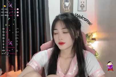 Media: A video of an Asian woman with long black hair, red lipstick, and pink shirt, sitting in a pink chair, eyes closed, in a dimly lit room with black curtains and a \"Welcome!\" sign.