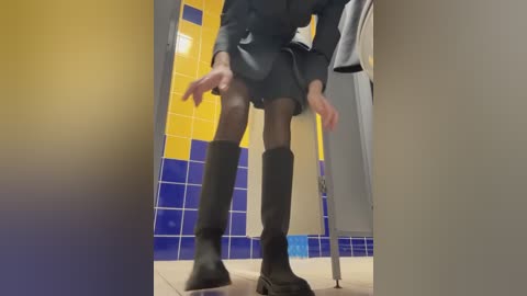 Media: Video of a person wearing a dark gray suit, black knee-high boots, and black stockings, standing in a public restroom with yellow and blue tiled walls.