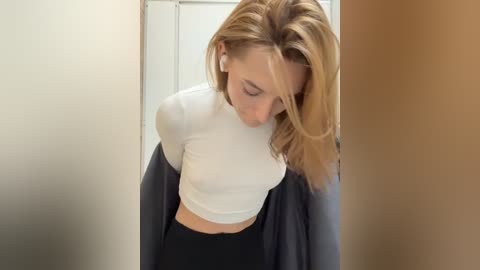 Media: Video of a blonde woman in a white crop top and black leggings, leaning slightly forward, looking down, in a dimly lit hallway.