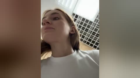 Media: A video of a young, light-skinned woman with straight, shoulder-length blonde hair, wearing a white top, standing in a modern bathroom with a white tiled wall and a grid-patterned glass window.