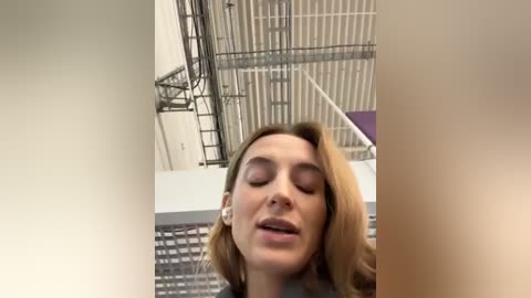 Media: Video of a young woman with shoulder-length blonde hair, closed eyes, and a serene expression, standing in a modern, industrial space with white metal beams and a purple wall.