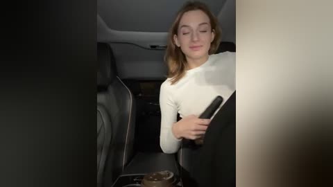 Media: A video of a young Caucasian woman with light brown hair, wearing a white sweater, sitting in a black car, reading a smartphone, with dim lighting.