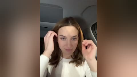 Media: Video of a young Caucasian woman with shoulder-length brown hair, fair skin, and light makeup, adjusting her hair in a dimly lit room with gray walls and a large round mirror.