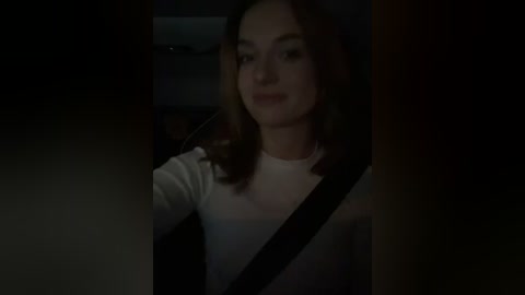 Media: A dimly lit video of a young woman with shoulder-length brown hair, wearing a white shirt, sitting in a car with a black seatbelt.