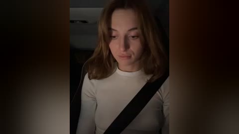 Media: Video of a Caucasian woman with shoulder-length light brown hair, wearing a white long-sleeve top, seated in a car, with a dark seatbelt across her chest, dimly lit interior background.