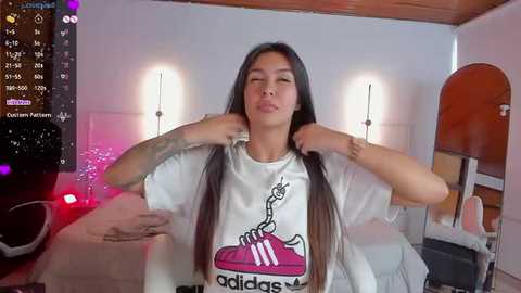 Media: Video of a young Asian woman with long black hair, wearing a white Adidas t-shirt, adjusting her shirt in a modern, brightly-lit bedroom.