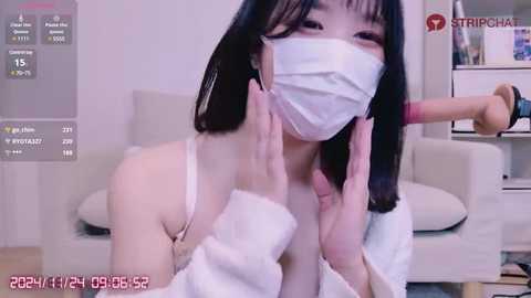 Media: A video of an East Asian woman with black hair, wearing a white face mask and a white top, kneeling in a living room.